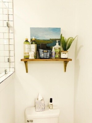 A bathroom with a white toilet, a shelf with toiletries, a plant, and a painting of a landscape. - Charleston Guest House- Easy parking! Quiet, safe neighborhood! Close to Dwntn! (Charleston)