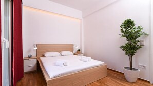 A neatly made bed with white linens and two pillows, a bedside table with a lamp, a small potted plant, and a wooden floor. - 45m2 homm Renovated Flat in Neos Kosmos Roumpesi str (Athens)