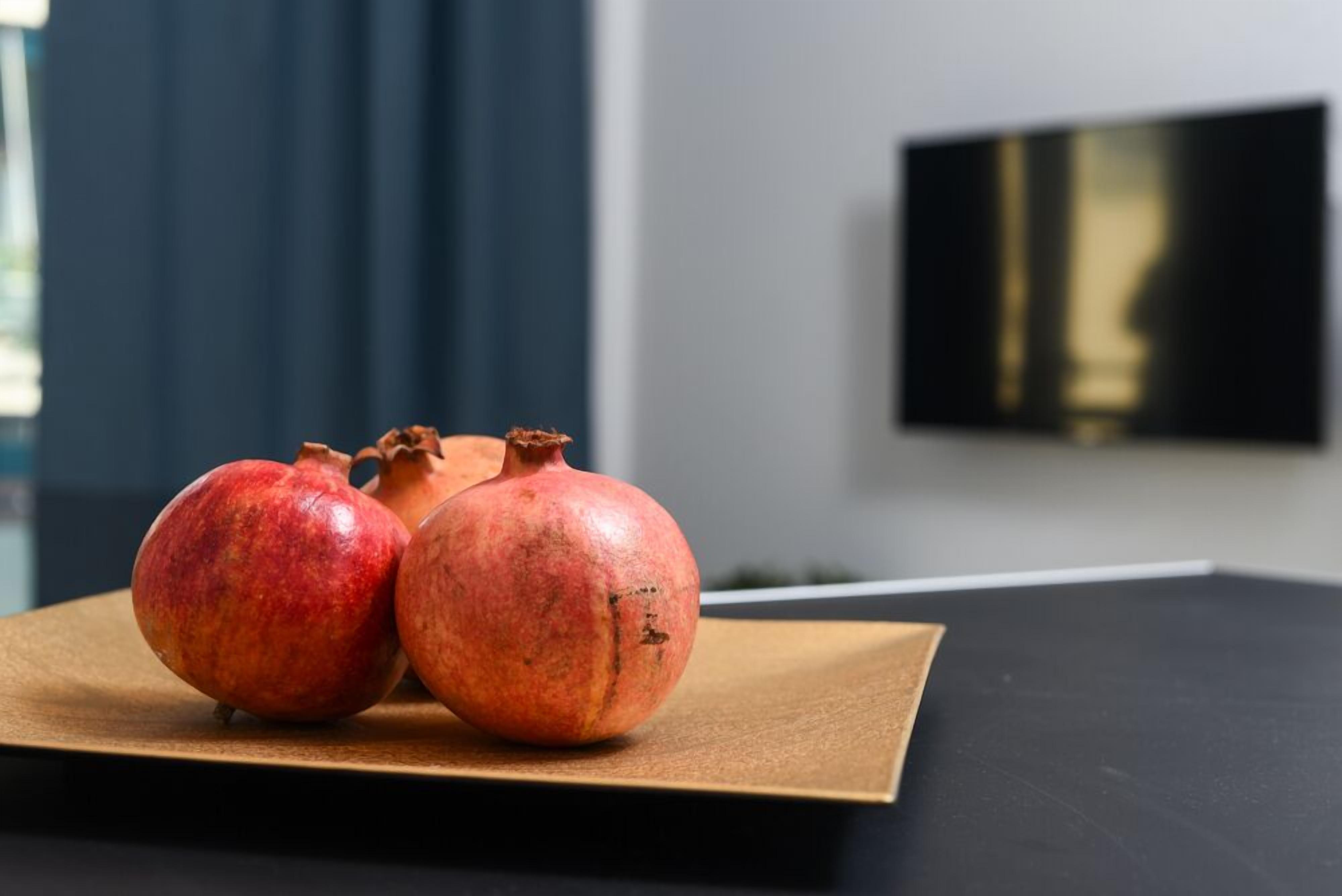 Zwei Granatäpfel auf einer braunen, rechteckigen Oberfläche mit schwarzem Hintergrund. An der Wand ist ein Fernseher angebracht.