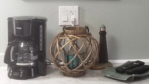 Un tableau représentant un phare au coucher du soleil, accroché au mur, une machine à café, un panier en osier avec une bougie, et une télécommande posée sur un comptoir.
