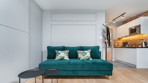 A modern living room with a teal sofa, two black side tables, and a kitchen area with white cabinets and a brick backsplash. - Barbakan Residence Old Town (Kraków)
