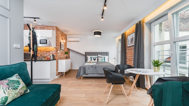 A compact living space with a kitchenette, a bed with decorative pillows, a dining area with a table and chairs, and a view of the outside through a large window. - Barbakan Residence Old Town (Kraków)