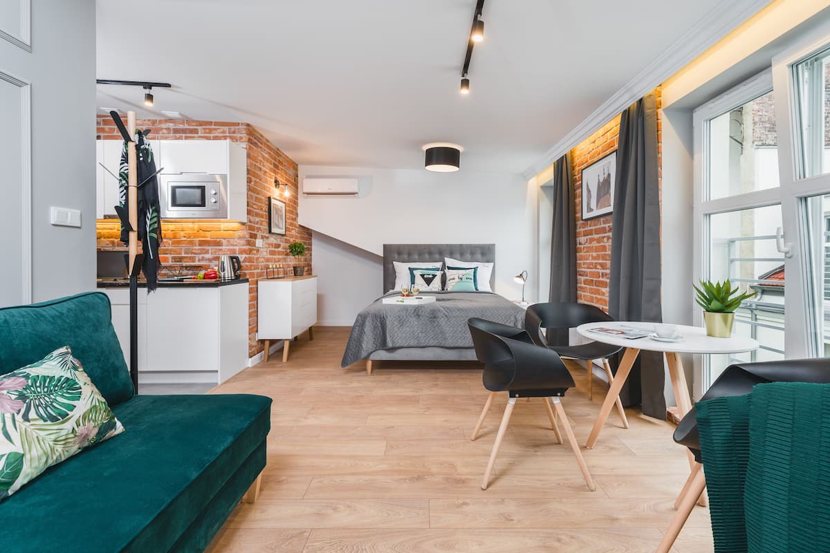 Un espacio habitable compacto con kitchenette, una cama con almohadones decorativos, zona de comedor con mesa y sillas, y vistas al exterior a través de un ventanal grande.