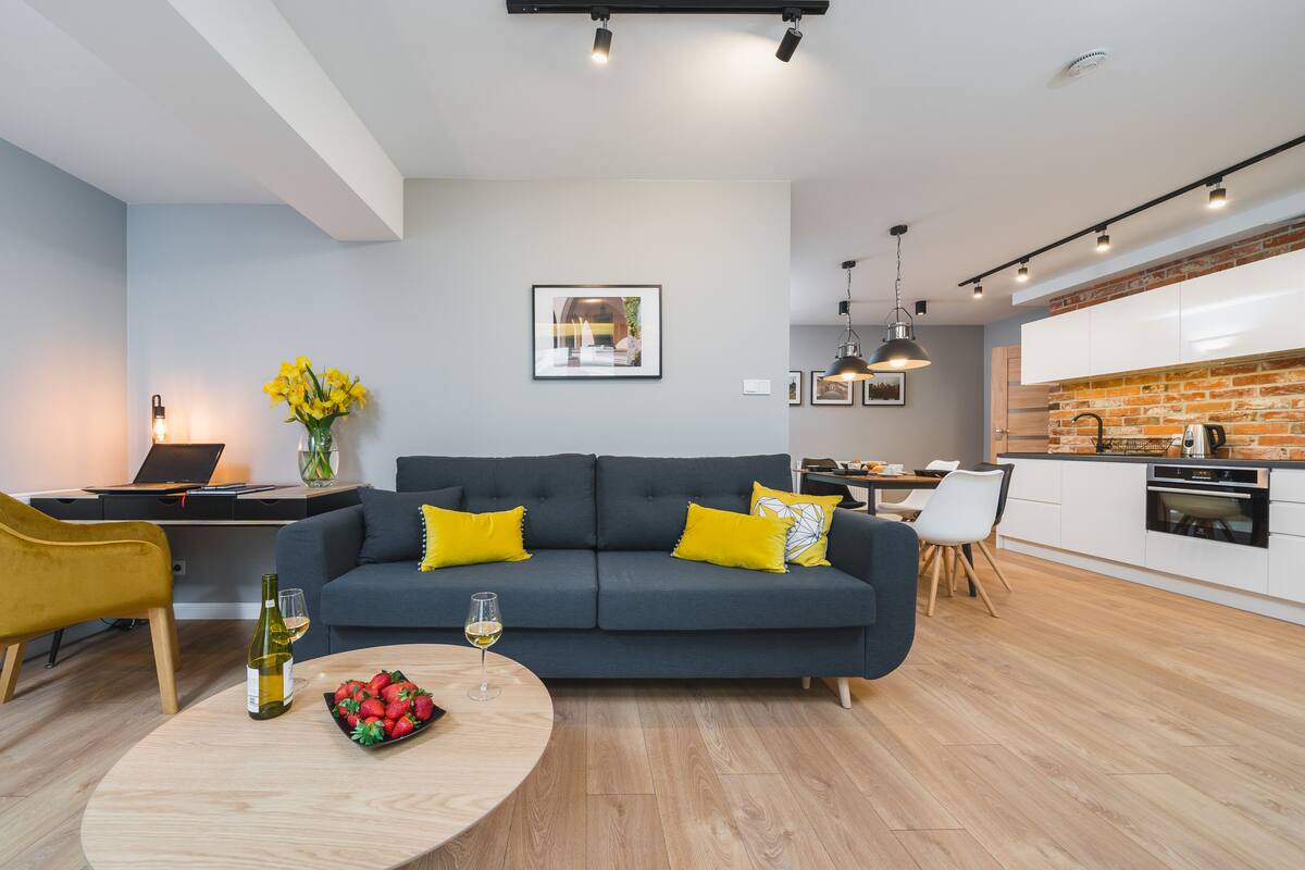 Una sala de estar moderna con un sofá gris oscuro, cojines amarillos, una mesa de centro de madera con fresas y vino, un portátil sobre un escritorio y una zona de comedor con un salpicadero de ladrillo.