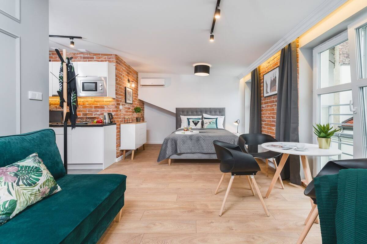 Un espacio habitable compacto con kitchenette, una cama con ropa de cama gris, un sofá verde, una mesa de comedor con sillas y un ventanal con cortinas.
