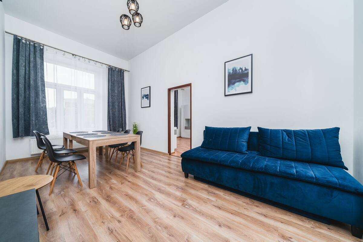 Una sala de estar moderna con piso de madera, un sofá azul, una mesa de comedor con sillas y un ventanal con cortinas.