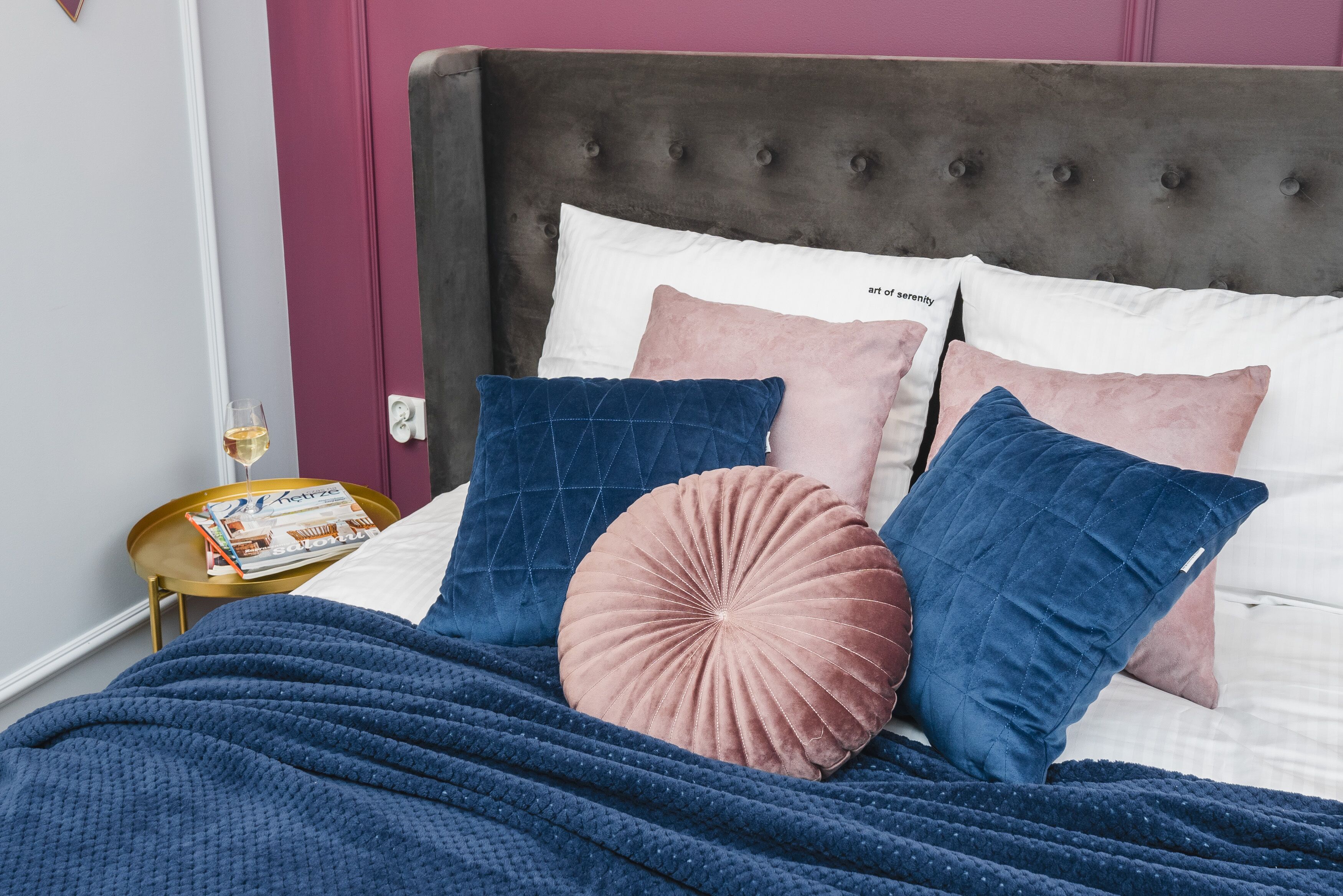 Ein ordentlich bezogenes Bett mit einer dunkelblauen Tagesdecke, rosa und blauen Kissen und einer Zeitschrift auf einem Nachttisch.