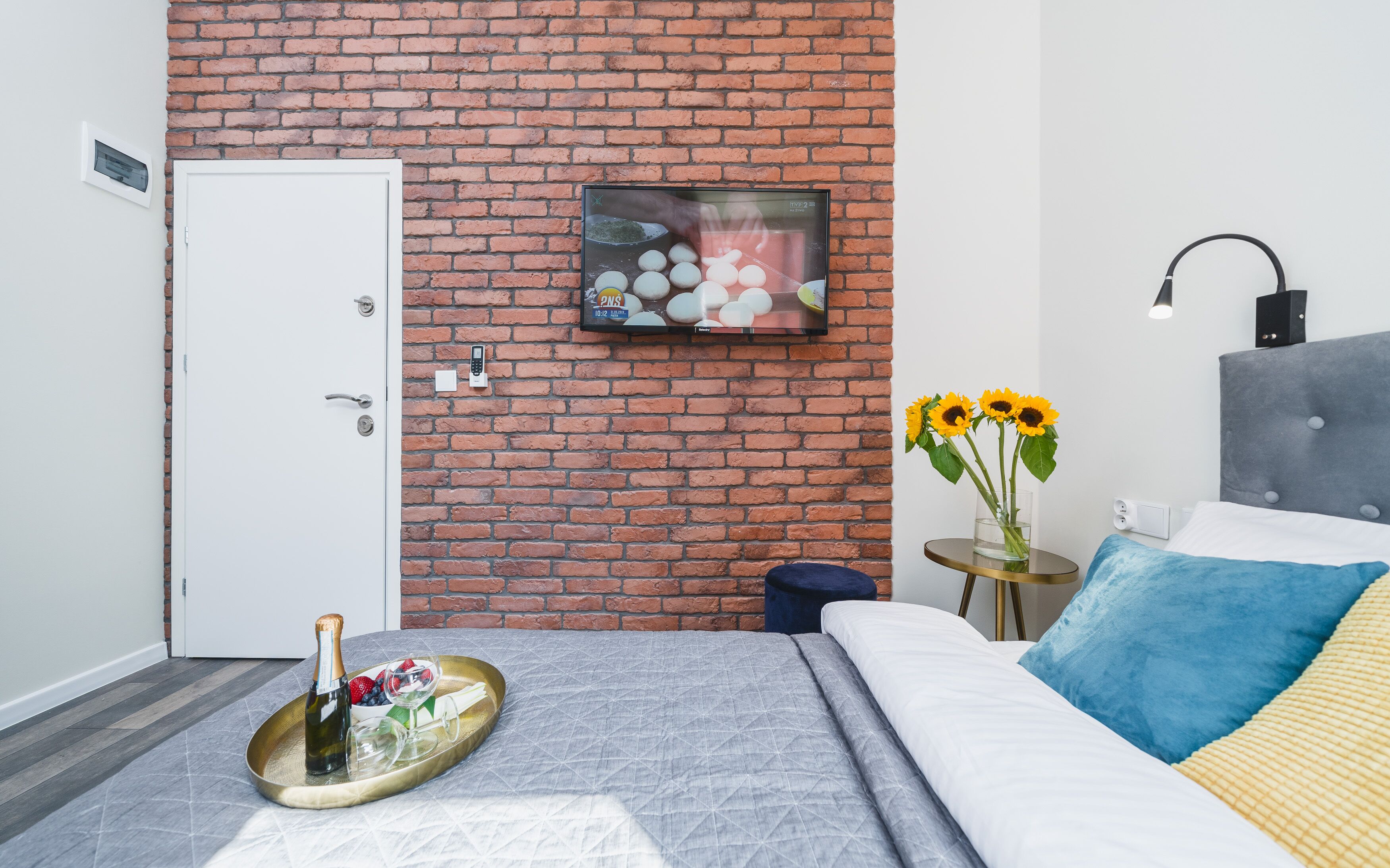 Una camera da letto moderna con una parete in mattoni, un letto con copripiumino grigio e blu, un comodino con un vaso di girasoli e una televisione a parete.