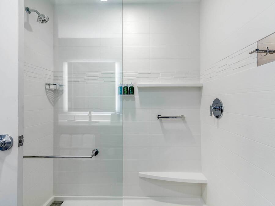 A modern bathroom with white tiles, a glass shower enclosure, a vanity with a mirror, and a small window.
