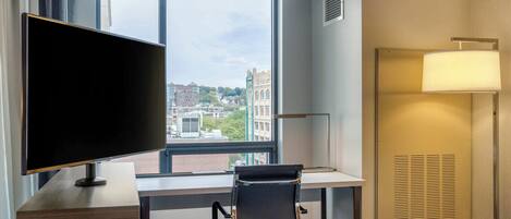 A hotel room with a large window, a desk with a chair, a flat-screen TV, a bed, and a wall-mounted lamp.