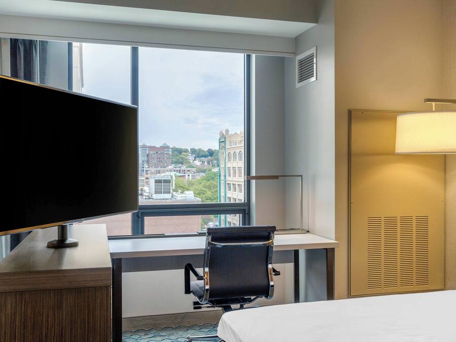 A hotel room with a large window, a desk with a chair, a flat-screen TV, a bed, and a wall-mounted lamp.