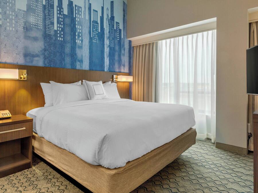 A hotel room with a large bed, a nightstand with a telephone, a flat-screen TV, and a cityscape painting on the wall.
