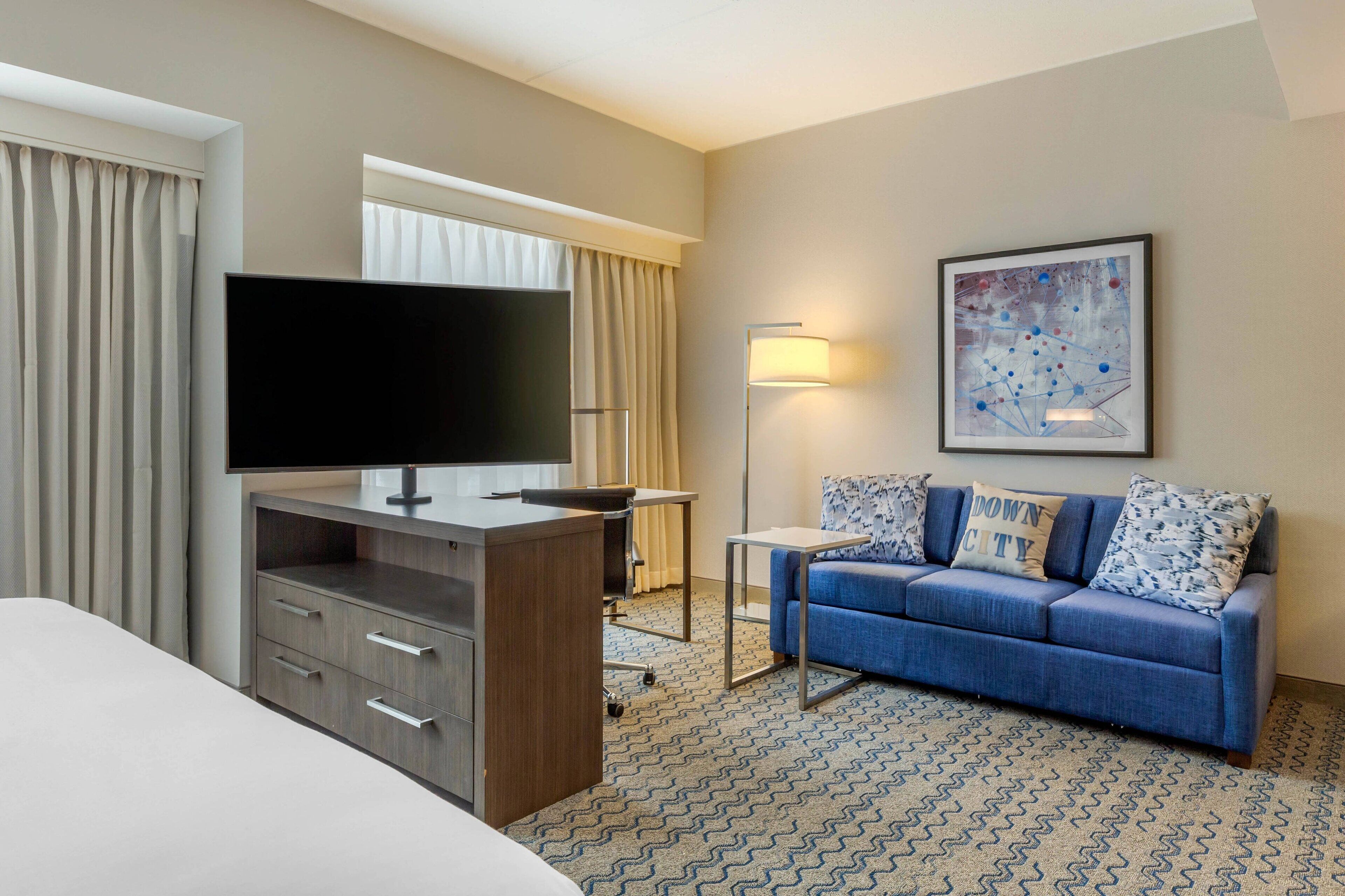 Habitación de hotel con cama, escritorio con televisor de pantalla plana, sofá con cojines estampados, lámpara de pie y un cuadro abstracto enmarcado en la pared.