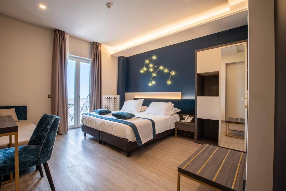 A modern hotel room with a large bed, a desk, a chair, and a balcony.