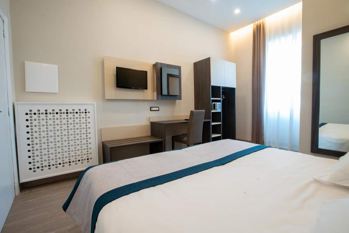 A hotel room with a bed, a desk, a chair, a TV mounted on the wall, and a wardrobe.