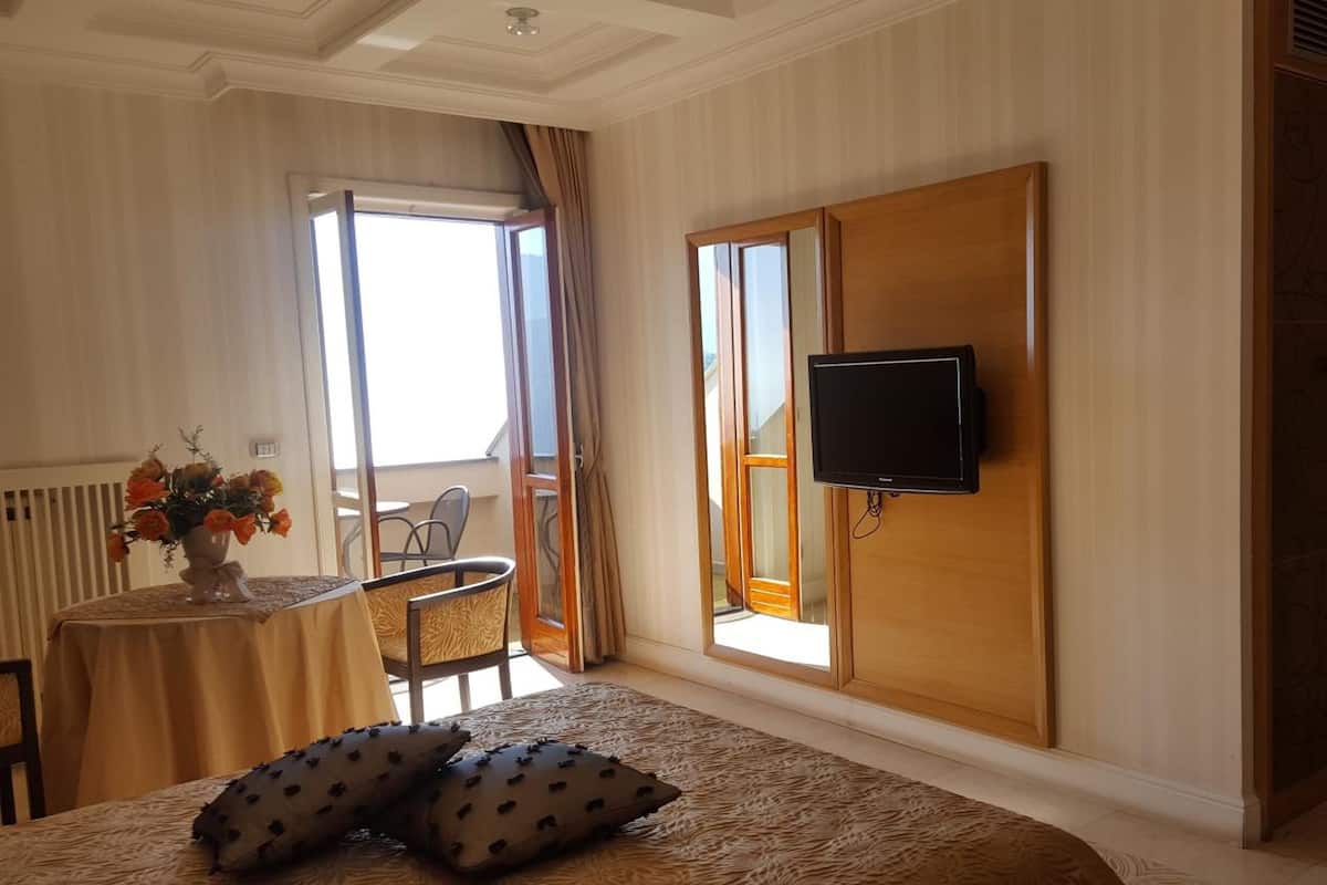 A hotel room with a bed, a TV mounted on the wall, a balcony with a chair, and a table with a vase of flowers.