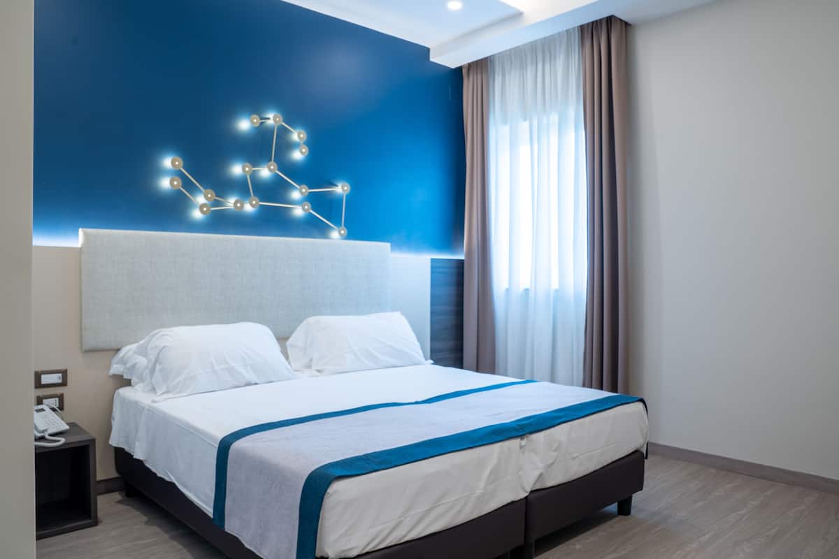 A modern hotel room with a large bed, a headboard with integrated lighting, a bedside telephone, and a window with curtains.