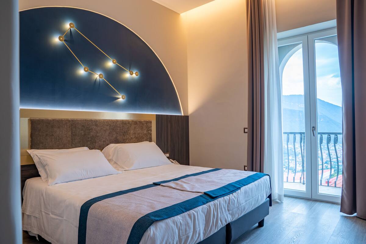 A modern hotel room with a large bed, a curved wall light fixture, and a balcony with a view.
