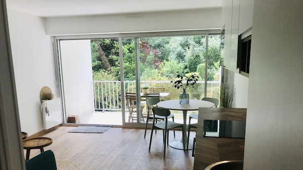 Una cocina moderna con encimera de madera, zona de comedor con mesa redonda y sillas, y puerta corredera de cristal que da a un balcón con vistas a zonas verdes.
