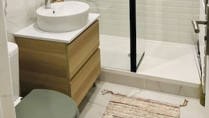 Un baño moderno con un lavamanos blanco, tocador de madera, espejo redondo, cabina de ducha con paredes de vidrio y un elemento vertical negro.