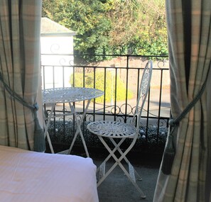 A balcony with a white metal table and chair, a white bed, and a view of a garden and street.