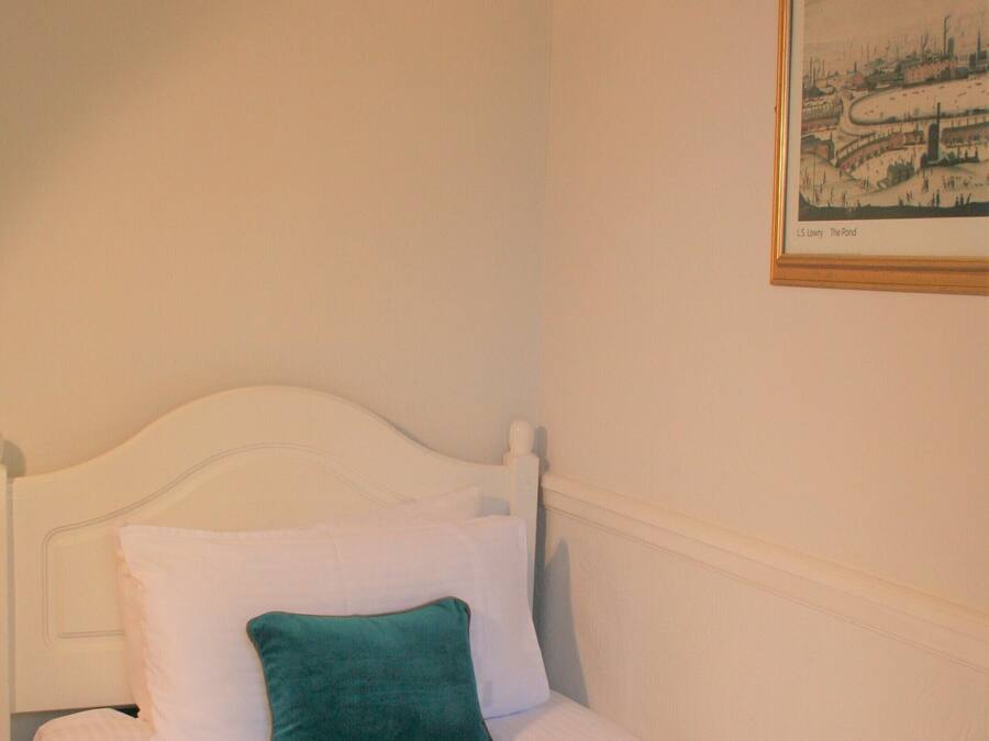 Una cama bien hecha con sábanas blancas y un cojín verde azulado, una lámpara de pared y un cuadro en la pared.