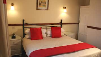 Ein ordentlich gemachtes Bett mit roter Decke und Kissen, ein Nachttisch mit Lampe sowie ein gerahmtes Bild an der Wand.