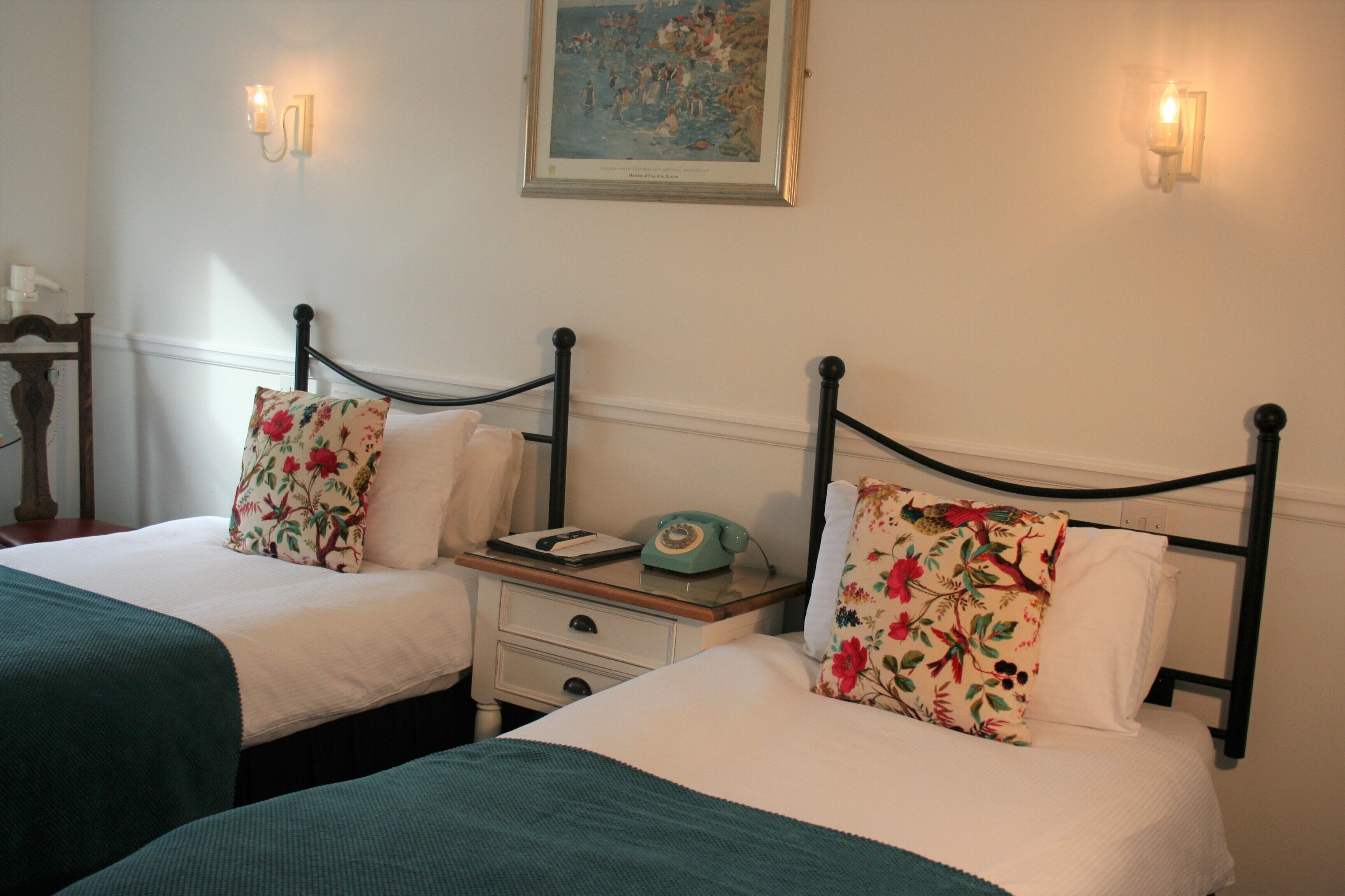 Une chambre d’hôtel avec deux lits simples, chacun agrémenté d’oreillers à motifs floraux et d’une couverture verte. Une table de chevet avec un téléphone d’époque se trouve entre les deux lits.