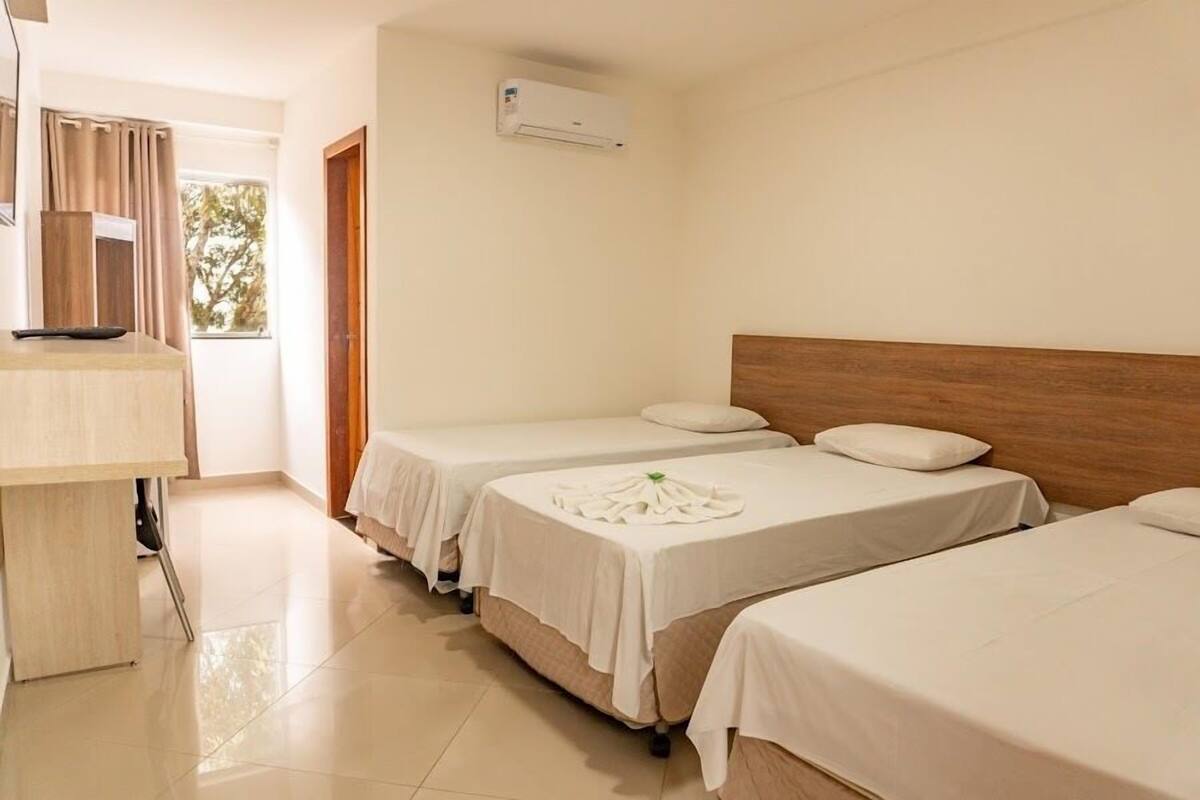 A hotel room with two beds, a desk, an air conditioning unit, and a view of trees through the window.