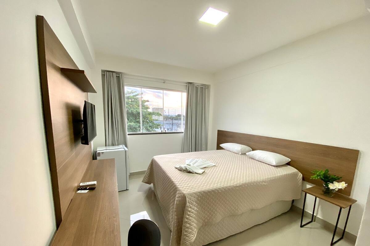 A hotel room with a bed, a desk, a chair, a TV, a window with curtains, and a small table with a plant.