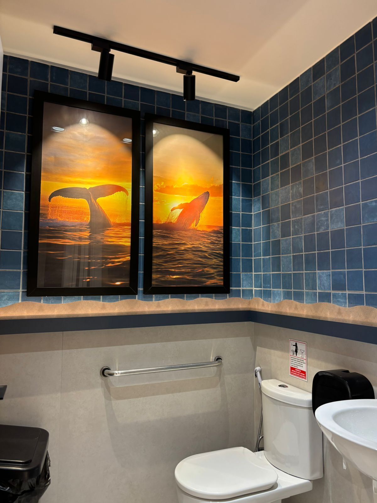 A bathroom with a toilet, bidet, and a sink. There are two framed pictures of whales with their tails above the water.