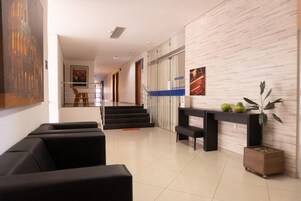 Um lobby moderno com escadaria, assentos pretos, balcão de recepção e uma planta em vaso.