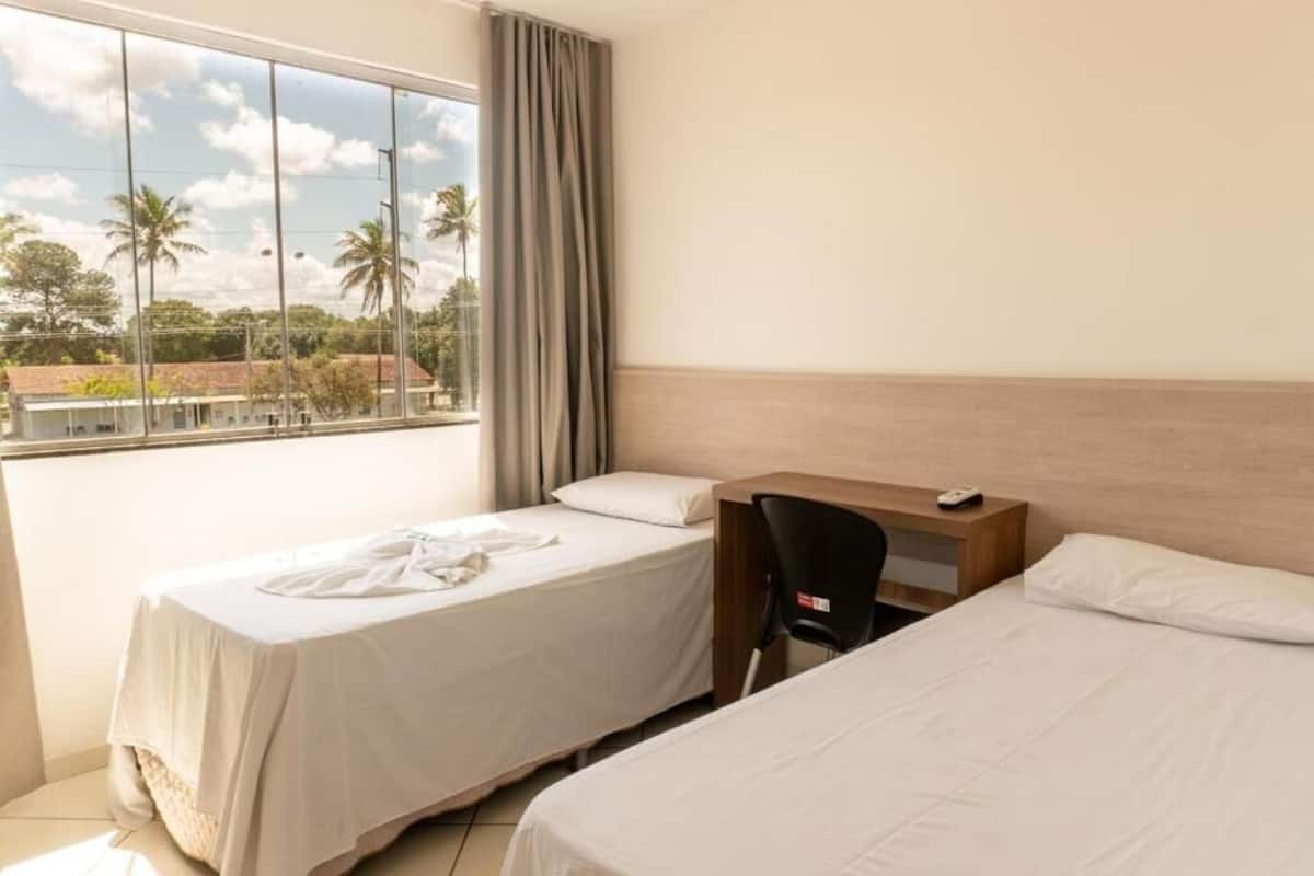 A hotel room with two beds, a desk, a chair, and a large window with a view of palm trees and buildings.