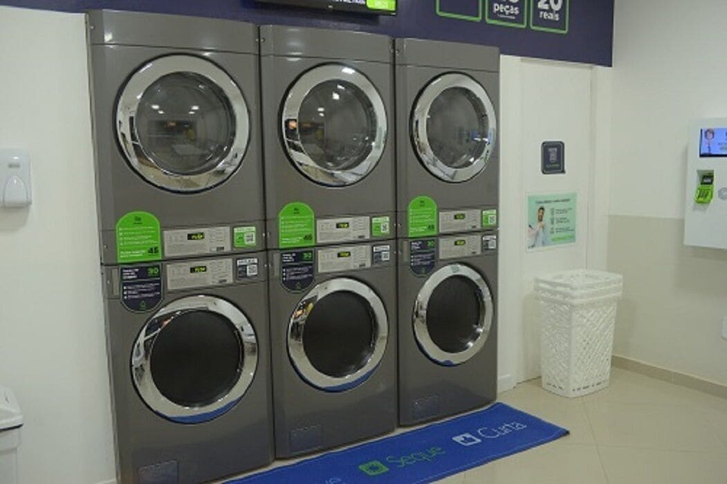 A row of six front-loading washing machines with control panels, a white trash bin, and a blue mat on the floor.