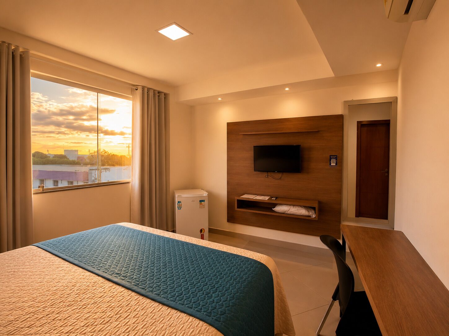 A hotel room with a bed, a desk, a television mounted on the wall, and a window with a view of buildings.