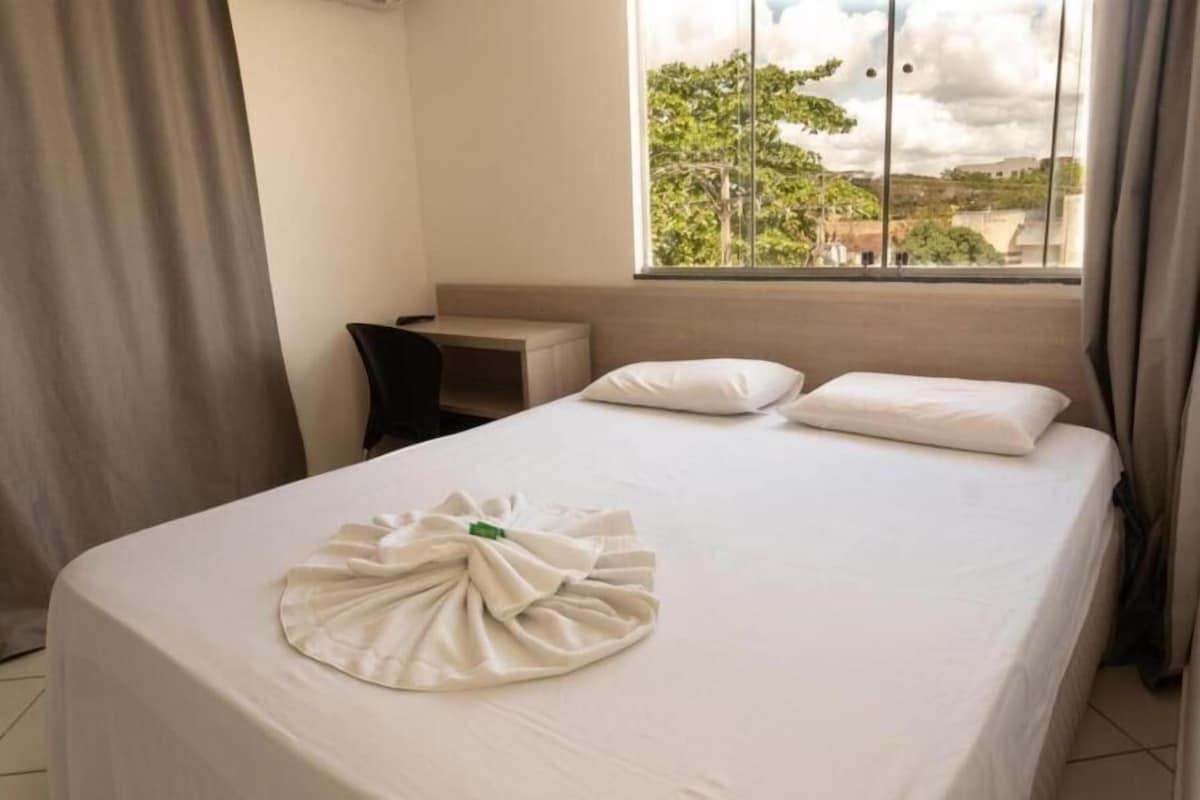 Habitación de hotel con cama, escritorio y ventana con vistas a árboles y edificios.