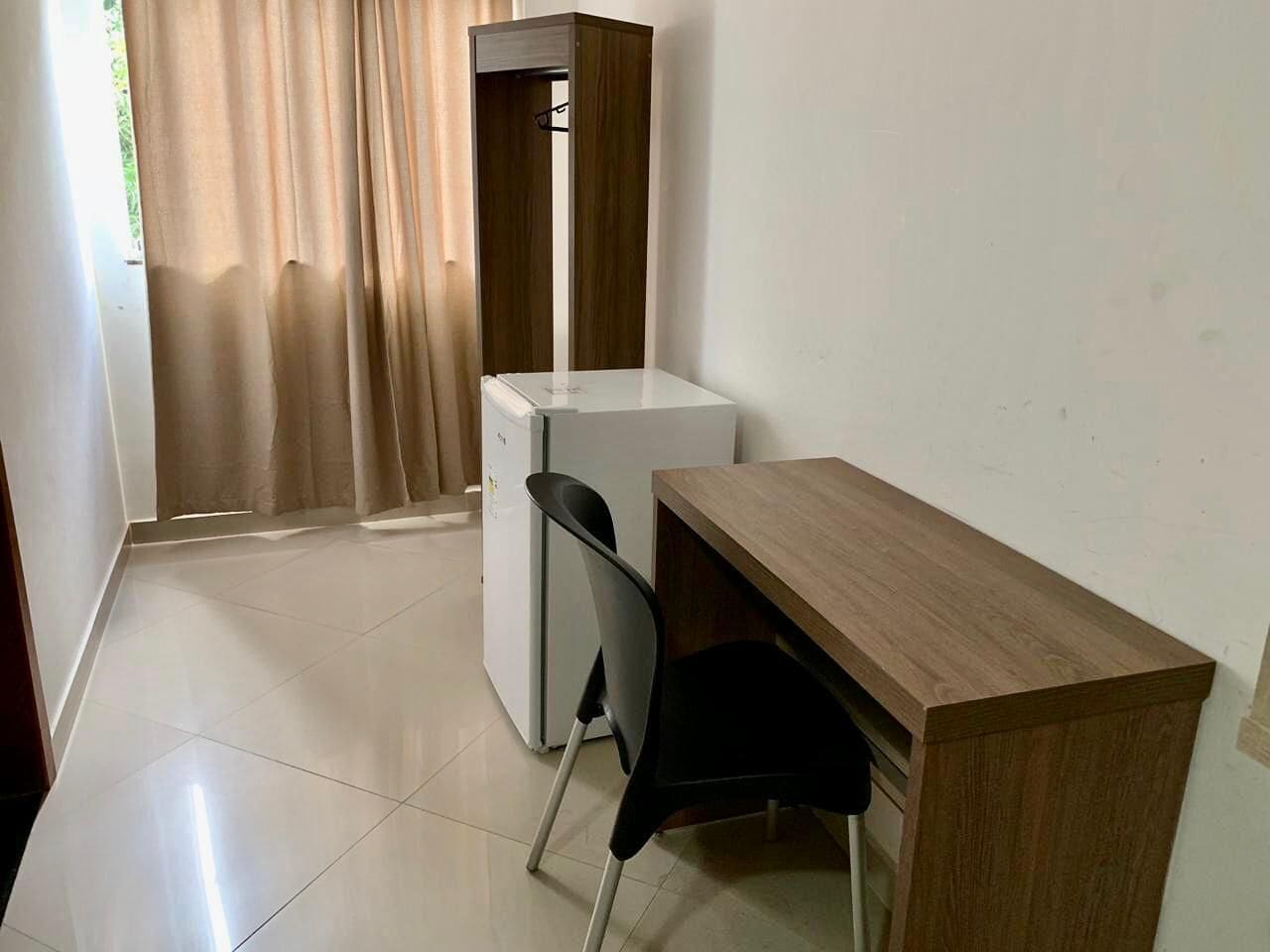 Quarto pequeno e limpo, com uma escrivaninha de madeira, uma cadeira e um mini-frigorífico branco.