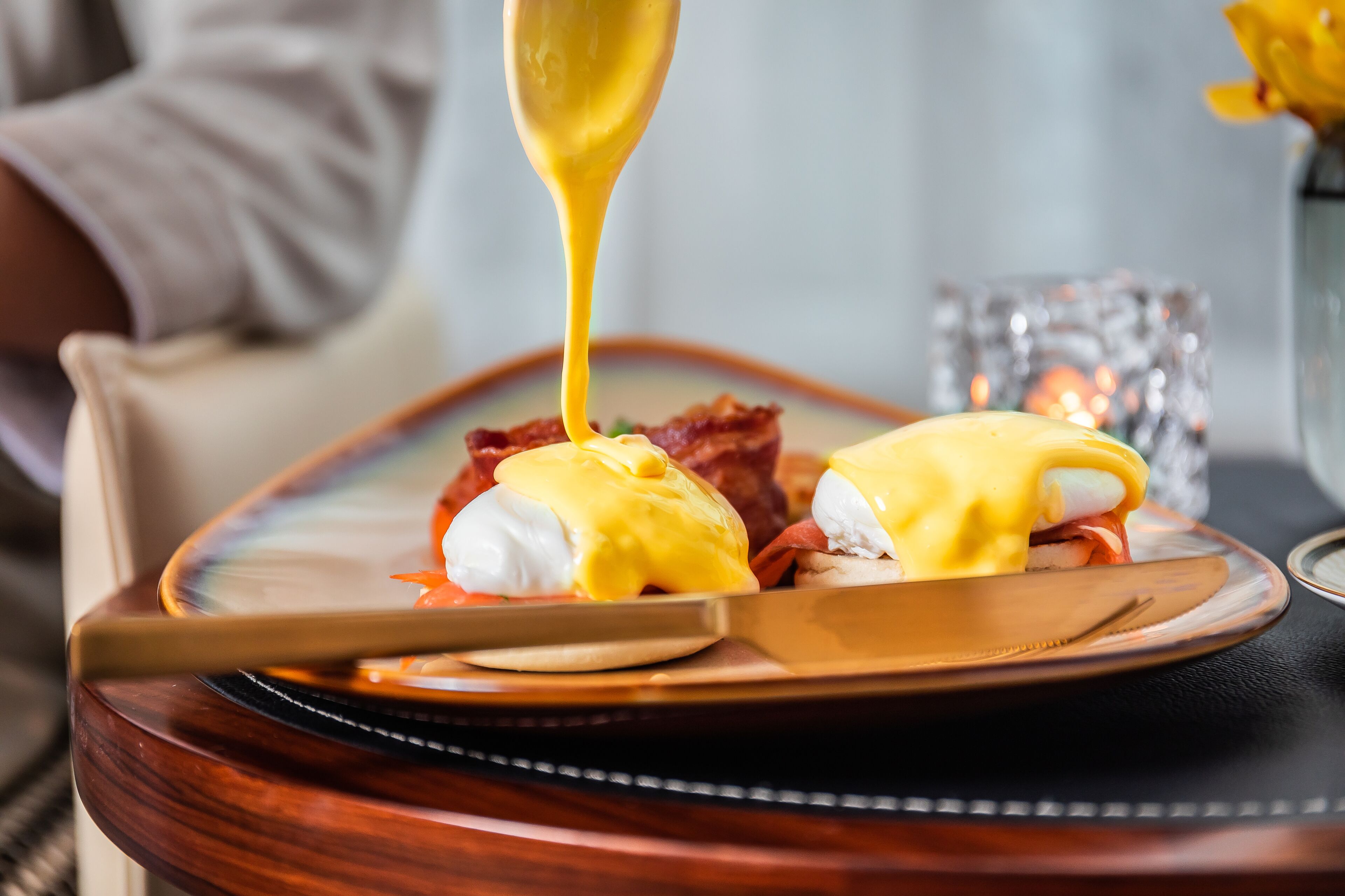 A plate with eggs benedict and bacon, a knife, and a spoon on a wooden table.