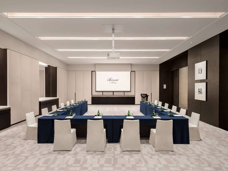 A conference room with a long table, white chairs, a projector screen, and a dark-colored reception desk.