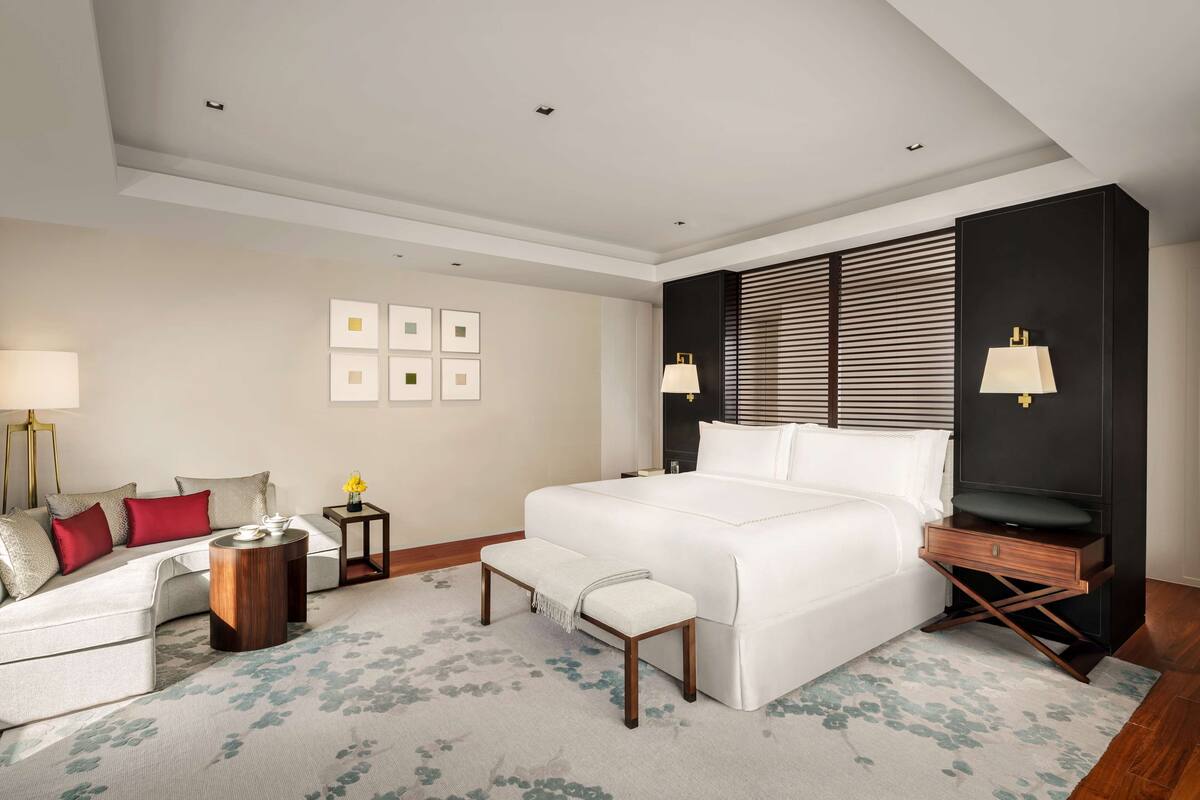 A modern hotel room with a large bed, a sofa, a bench, a nightstand, and a lamp.
