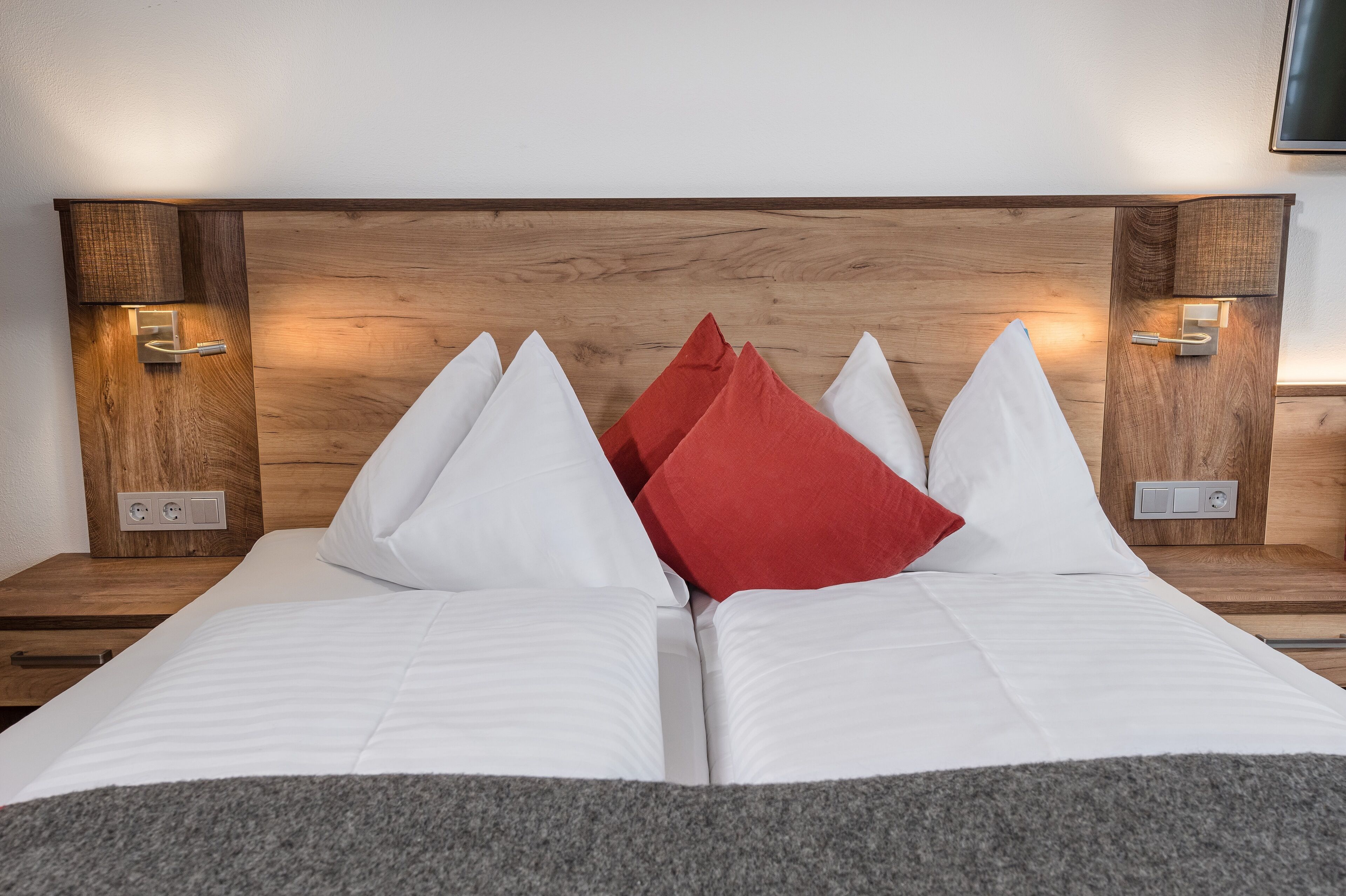 A double bed with white and red pillows, wooden headboard, bedside lamps, and power outlets.