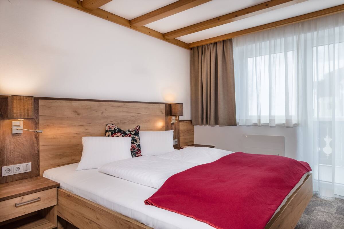 A hotel room with a wooden headboard, white bedding, a red throw blanket, a bedside table with a lamp, and a window with curtains.