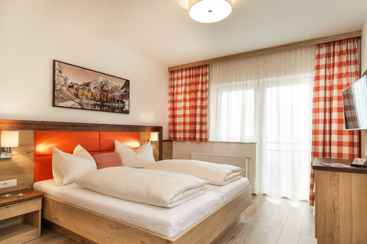 A modern hotel room with a large bed, wooden headboard, two bedside tables with lamps, a desk with a computer, and a window with checkered curtains.