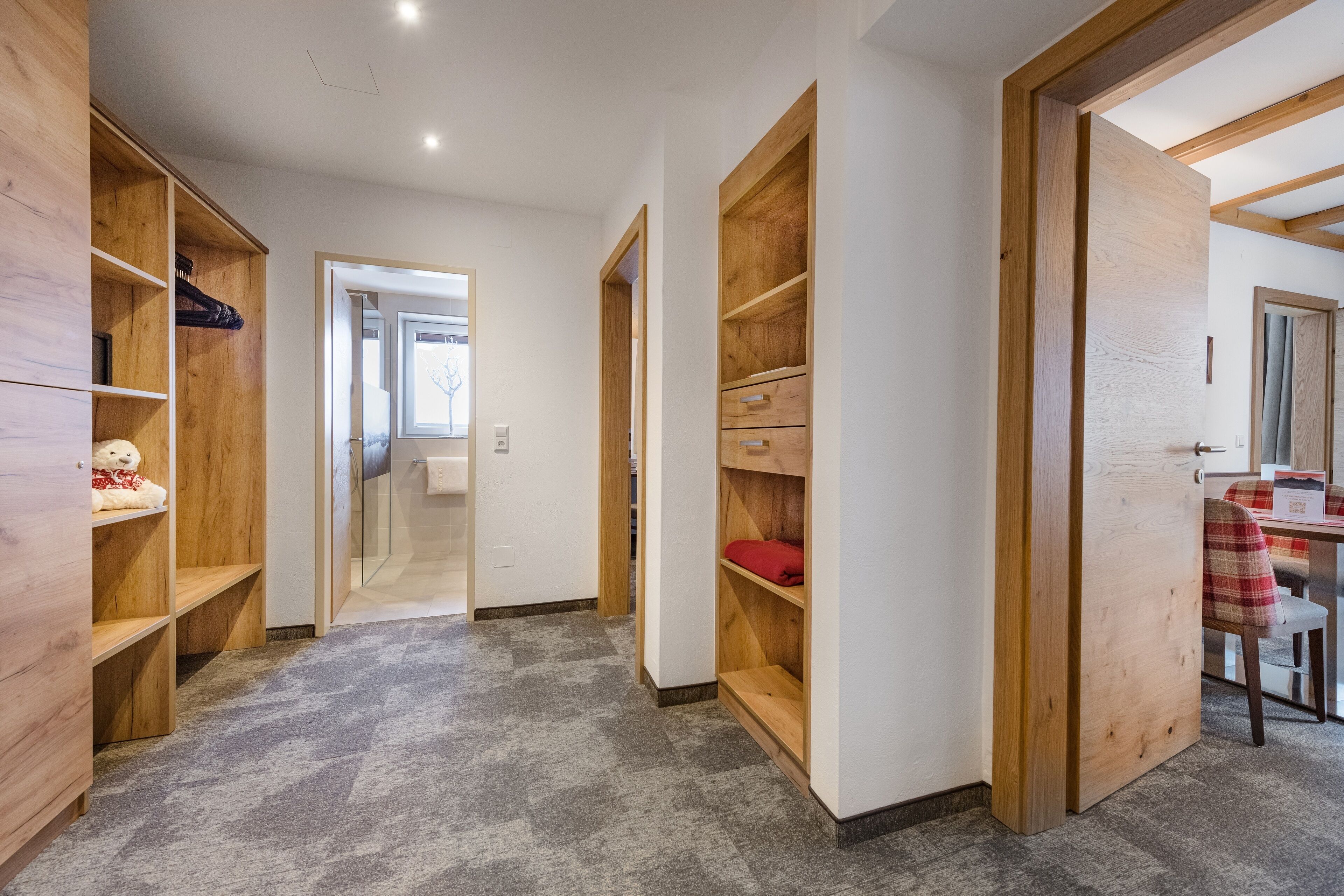 Un pasillo con estanterías de madera, una puerta de cristal que da al baño y una zona de comedor con mesa y sillas.