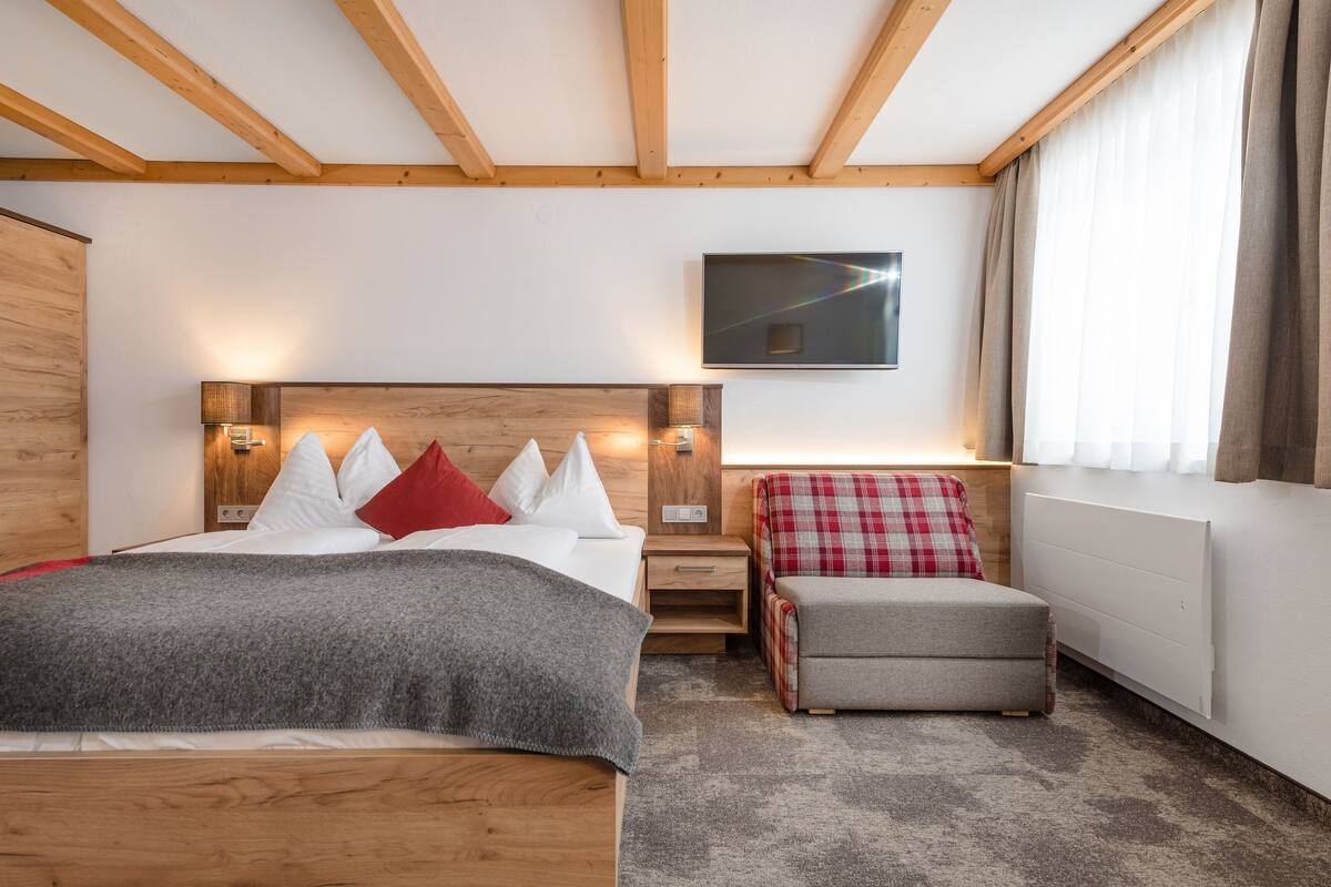 A hotel room with a wooden ceiling, a bed with a grey blanket, a red and white checkered chair, a flat-screen TV, and a window with curtains.