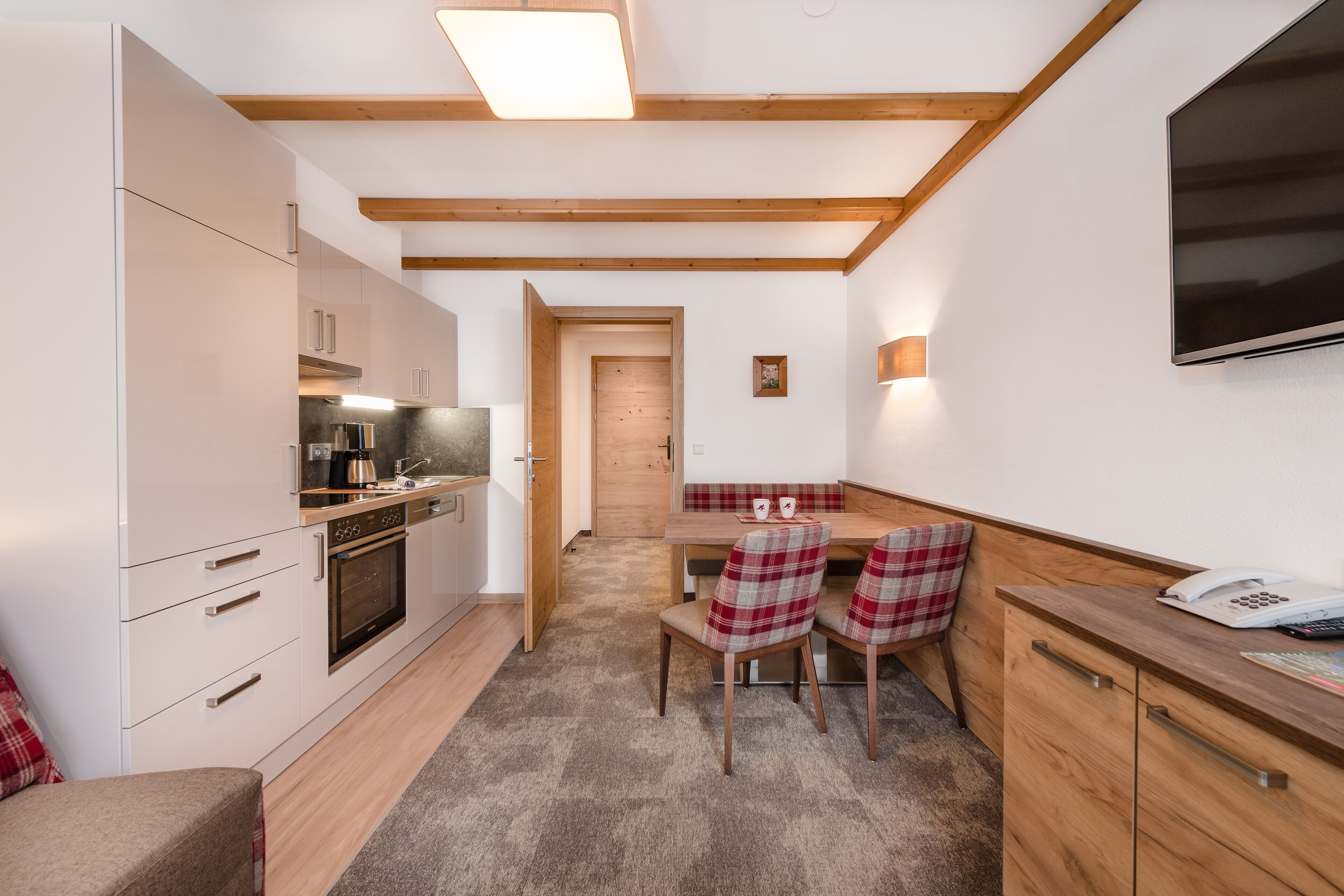 A compact kitchen with modern appliances, a dining area with plaid chairs, and a wooden ceiling.