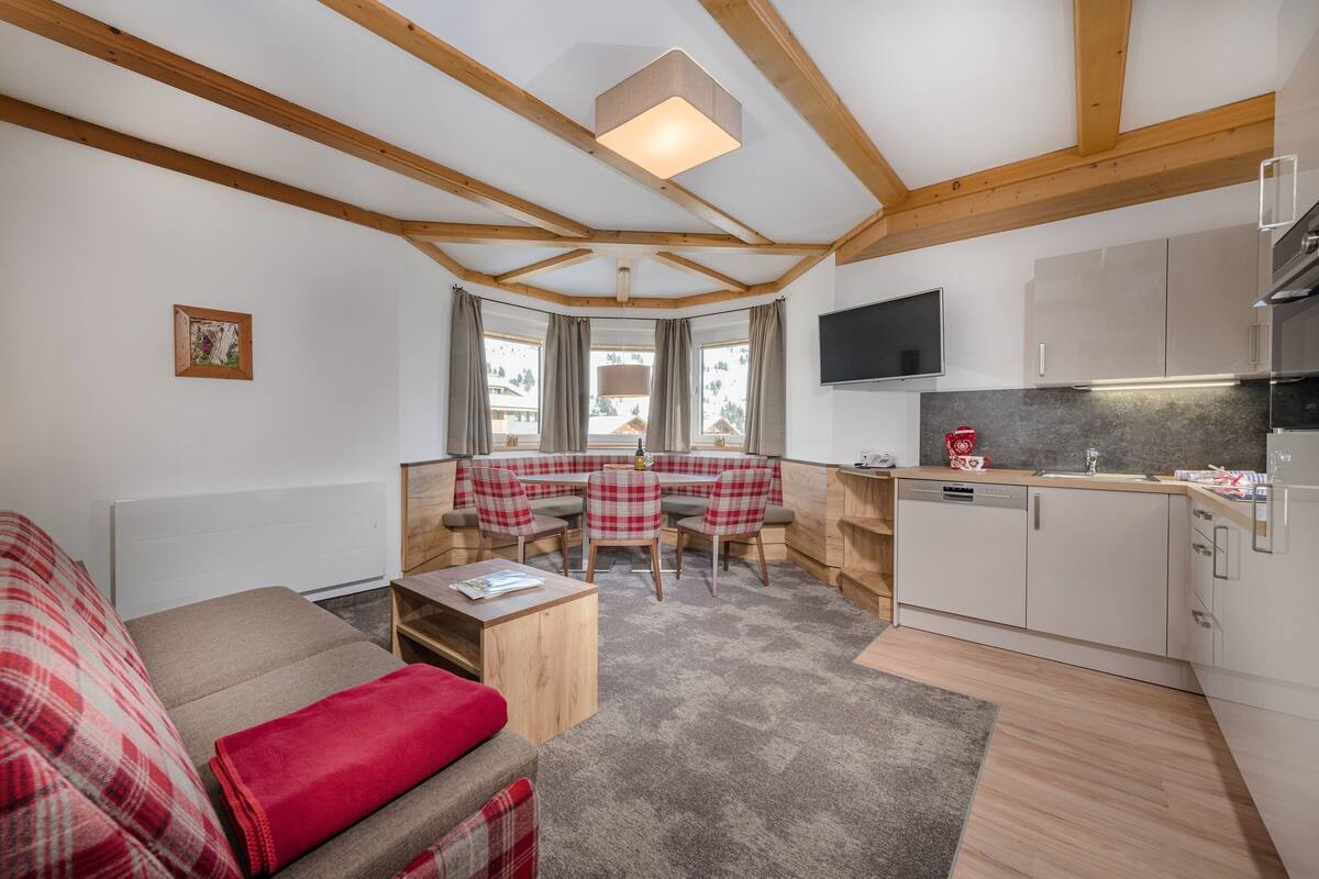 A cozy living area with a sofa, a small table, and a dining table with chairs. There is a kitchen area with cabinets and a TV.