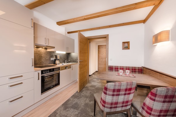 A modern kitchen with white cabinets, a built-in oven, and a wooden ceiling. There is a dining area with a wooden table and plaid chairs. - Apartments Garni Alpenrose (Tweng)