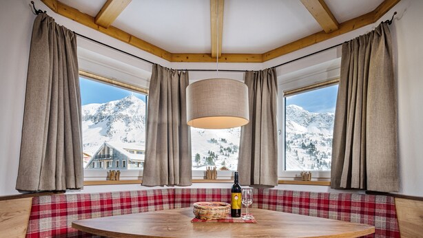 A cozy room with a round wooden table, plaid upholstered benches, and a view of snow-covered mountains through large windows. - Apartments  Garni Alpenrose (Tweng)
