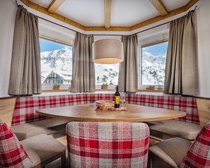 A cozy room with a round wooden table, plaid upholstered benches, and a view of snow-covered mountains through large windows. - Apartments Garni Alpenrose (Tweng)